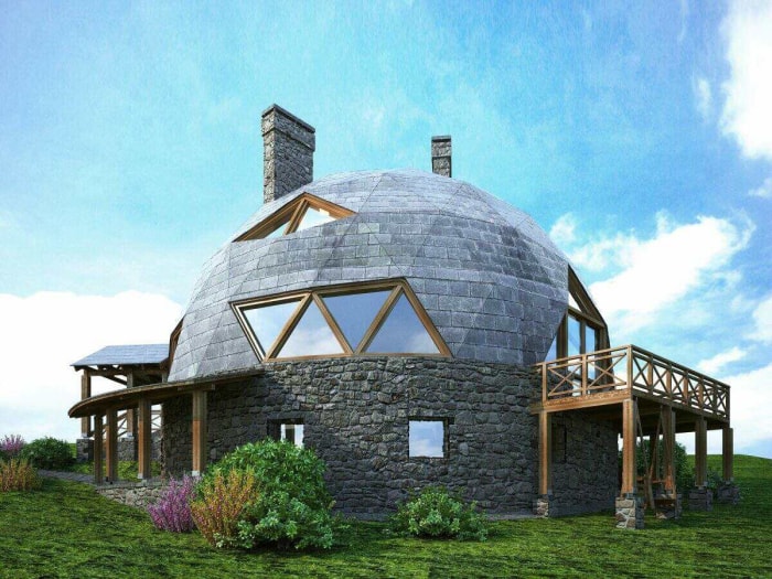 Une maison en forme de dôme avec des fenêtres triangulaires et une terrasse en bois enveloppante se dresse sur une colline verdoyante.