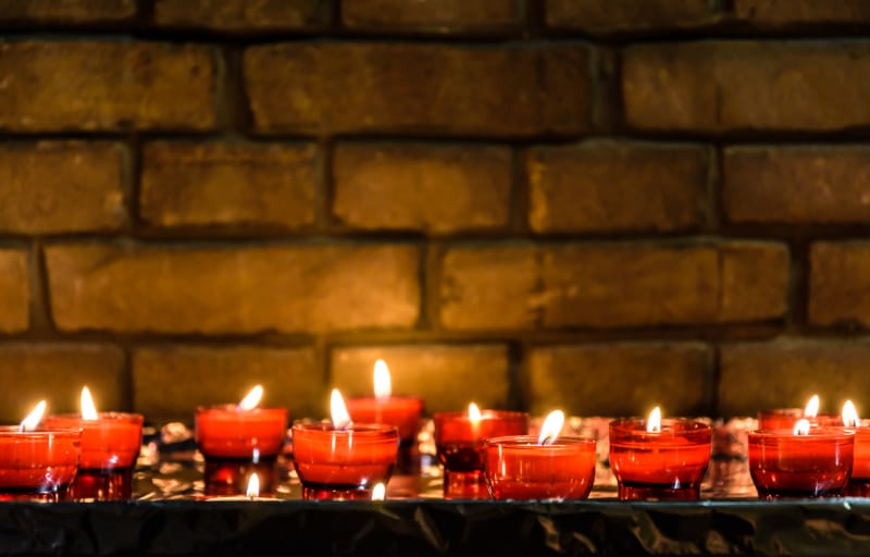 Petites bougies votives blanches allumées et placées dans des porte-verres rouges sur une table recouverte d'une feuille d'aluminium devant un mur de briques.