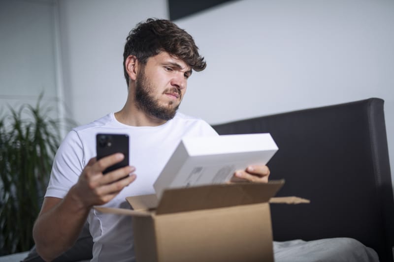 Un jeune homme regarde confusément le produit qui lui a été expédié, le comparant apparemment à l'article attendu sur son téléphone.