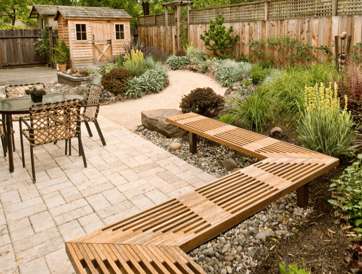 Un hangar en bois se trouve dans une cour arrière entourée de plantes avec un patio, une table et des chaises au premier plan.