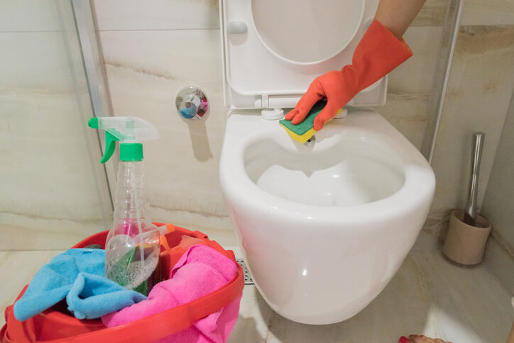 main dans un gant en organza utilisant une éponge pour nettoyer les toilettes avec un panier rose de produits de nettoyage à proximité dans une salle de bain beige