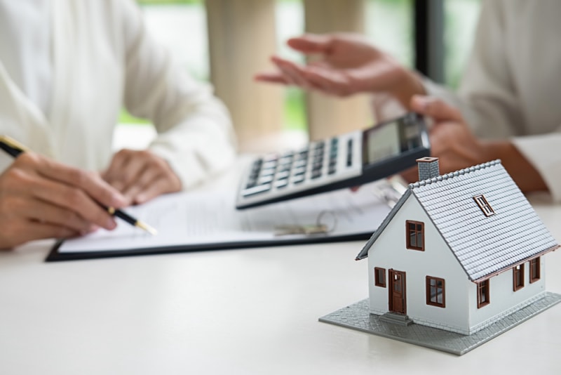 Une petite maquette de maison blanche est posée sur une table tandis que deux personnes discutent d'un document et de chiffres sur une calculatrice. 