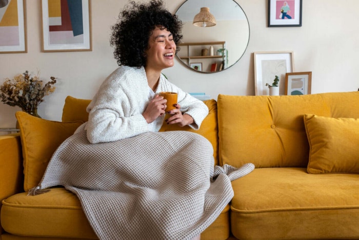 femme riant assise sur un canapé recouvert d'une couverture et tenant une tasse