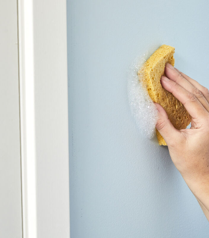 Une femme essuie un mur bleu avec une éponge jaune savonneuse.