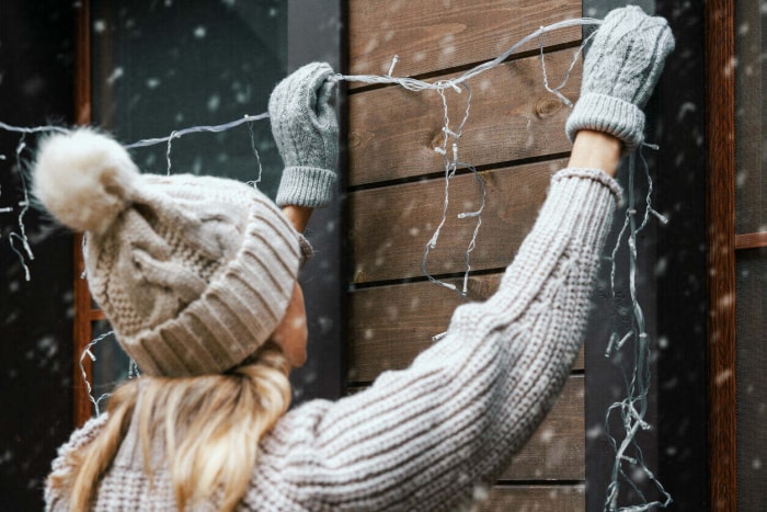 Une femme portant un chapeau d'hiver gris tient des guirlandes lumineuses contre le mur extérieur et les fenêtres pour les accrocher.