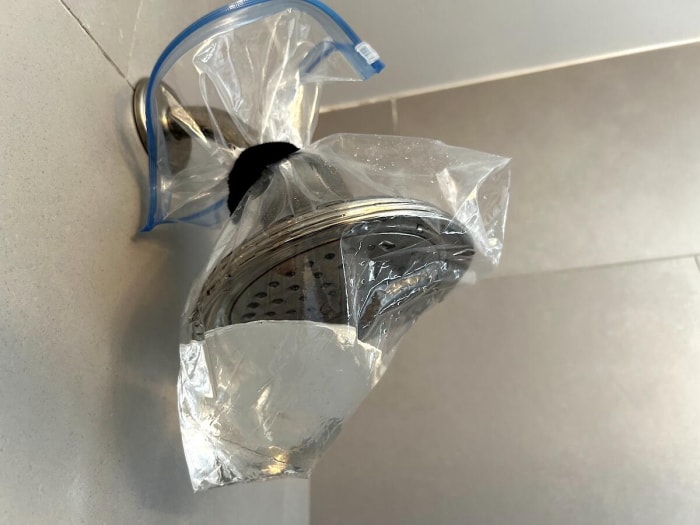 Un sac ziploc rempli de vinaigre noué autour d'une pomme de douche dans une douche carrelée grise.