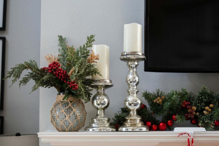 Un manteau de Noël blanc décoré avec des guirlandes de pin, des chandeliers en argent avec des bougies et une télévision.