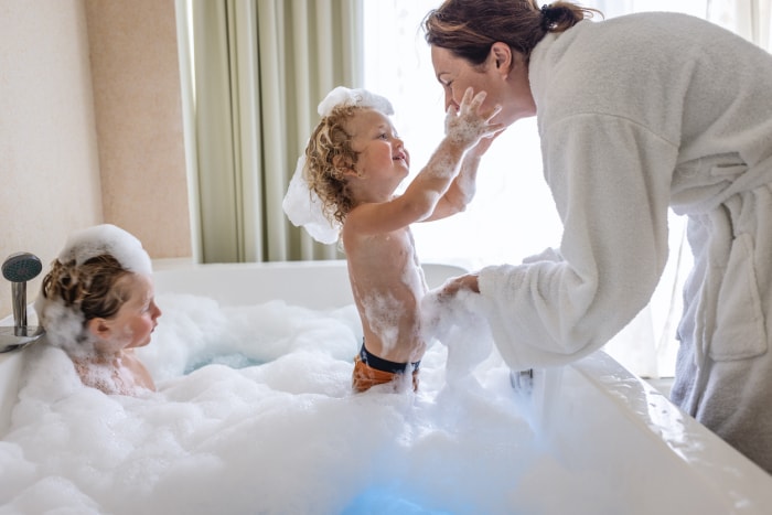 une femme en peignoir blanc se penche sur un bain moussant et joue avec un garçon tandis qu'un autre garçon regarde