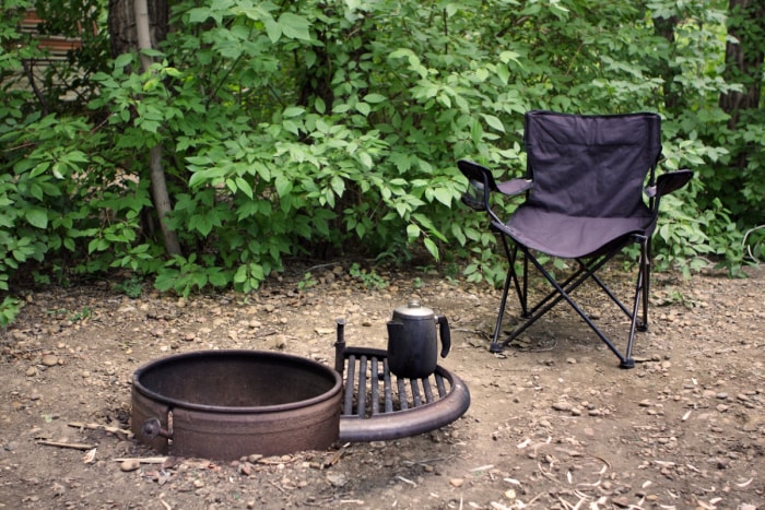 Une chaise de camping noire se trouve à côté d'un foyer vide avec une cafetière en métal noir posée dessus.