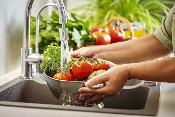 Gros plan des mains d'une femme tenant une passoire pleine de légumes frais sous l'eau courante dans l'évier de la cuisine.