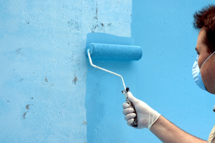 un homme portant un masque utilise un rouleau pour peindre un mur en bleu