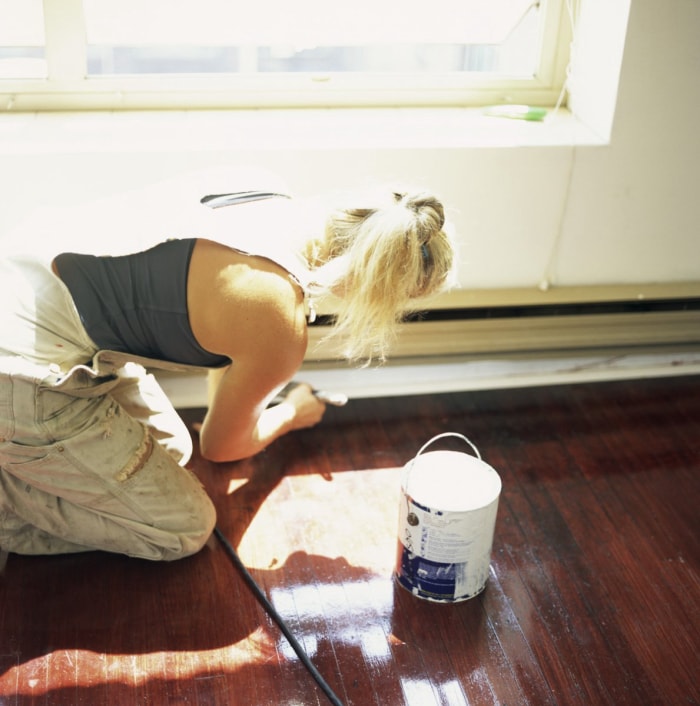 Une femme en salopette est accroupie sur le sol et peint les plinthes.