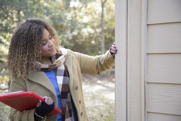 une femme souriante, vêtue d'un manteau et d'une écharpe, tenant un presse-papiers rouge, touche le revêtement beige de l'extérieur d'une maison pour l'évaluer.