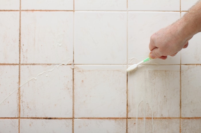 Une personne utilisant une brosse à dents pour nettoyer le coulis entre les carreaux muraux de douche sales.