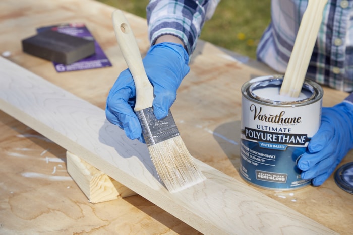 Une femme applique du polyuréthane sur une planche de bois avec un pinceau.