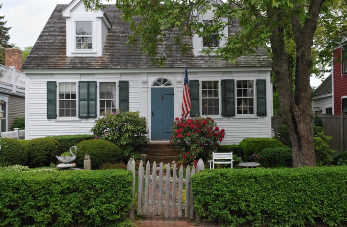 maison de style cottage blanc avec aménagement paysager, drapeau américain et haie pour la clôture avant