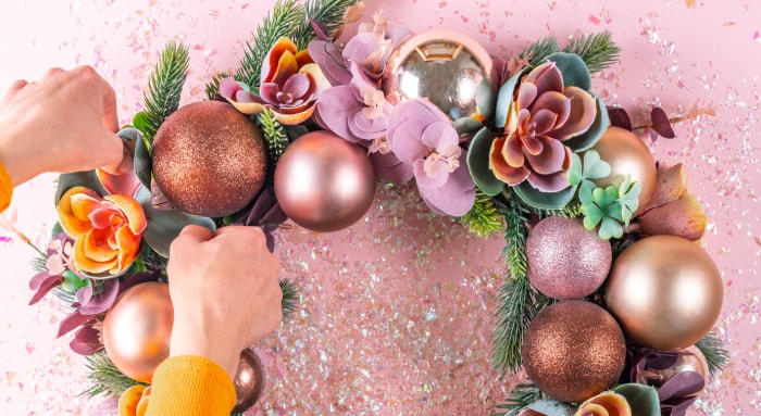 Des mains arrangent une fleur artificielle sur une couronne rose avec de la verdure et des décorations pour sapin de Noël.