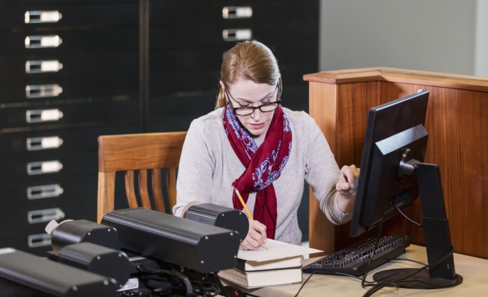 Une femme d'âge mûr dans une bibliothèque, utilisant un ordinateur pour consulter des documents historiques stockés sur des microfiches. Elle prend des notes.