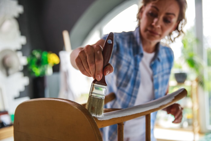 Focus sur le premier plan d'une femme mature peignant une chaise avec de la peinture blanche tout en apprenant un nouveau passe-temps pendant les jours de confinement à la maison.