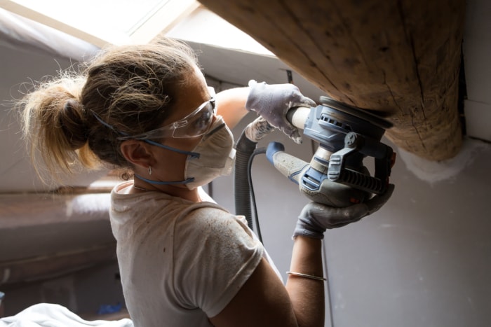 Une femme utilise une ponceuse sur une poutre en bois tout en portant un masque.