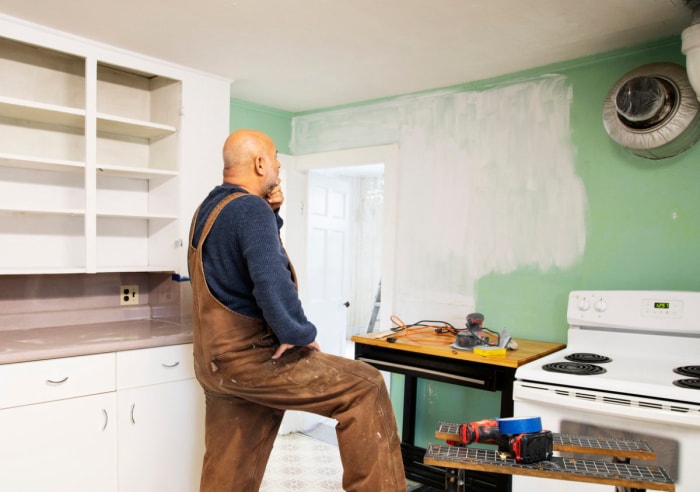 Un homme en salopette examine un mur partiellement peint dans une petite cuisine dépareillée.