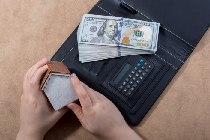 Gros plan sur les mains d'une personne tenant une petite maquette de maison à côté d'une calculatrice et d'un tas d'argent.