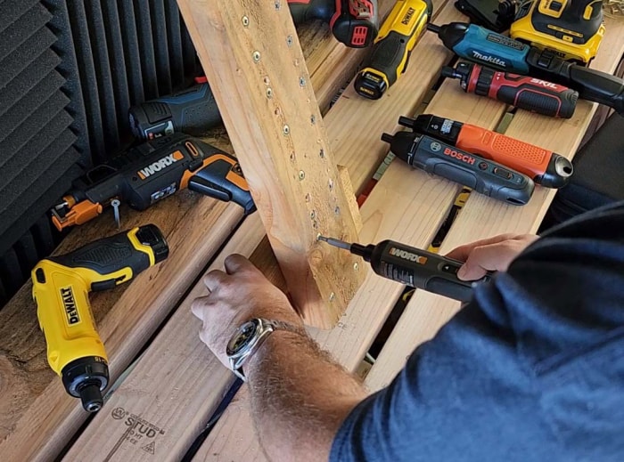 Une personne teste la meilleure option de tournevis sur un morceau de bois avec plusieurs autres tournevis électriques sur une table à l'arrière-plan.