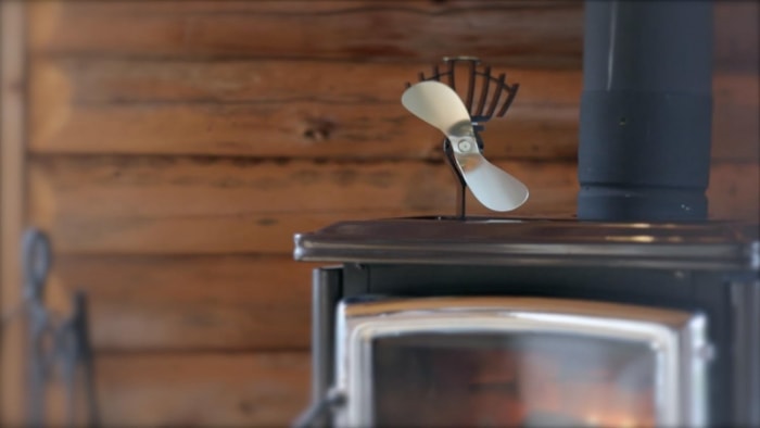 Le ventilateur de poêle à bois à pales en nickel Ecofan UltrAir sur le dessus d'un poêle à bois dans une pièce aux murs en bois.