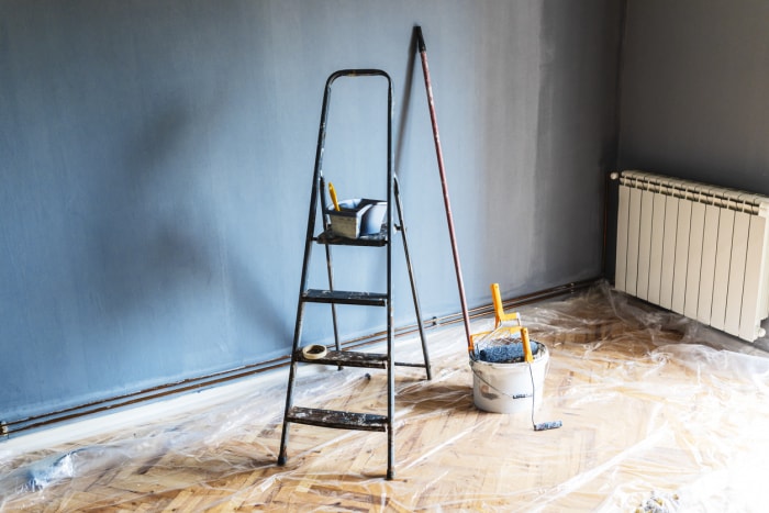 Outils de peinture et échelle dans une pièce vide préparée pour la rénovation avec une bâche en plastique recouvrant le sol.