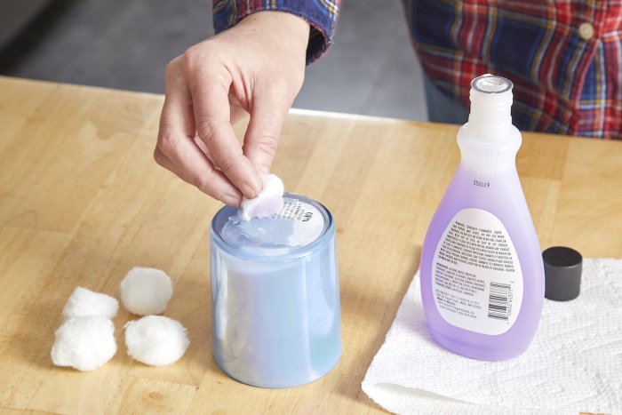 Une femme utilise du dissolvant pour vernis à ongles sur une boule de coton pour retirer un autocollant sur un pot de bougie.