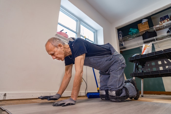 Un homme caucasien âgé en train de rénover un bâtiment à l'intérieur. Il est habillé en tenue de travail avec une salopette en jean et un t-shirt. Intérieur d'une maison privée en cours de rénovation, située en Amérique du Nord.