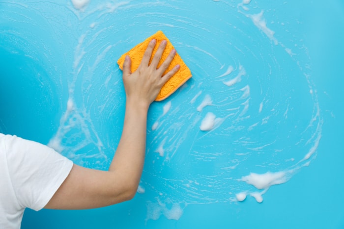 Une femme en chemise blanche lave les murs bleus avec un chiffon jaune et du savon à vaisselle.