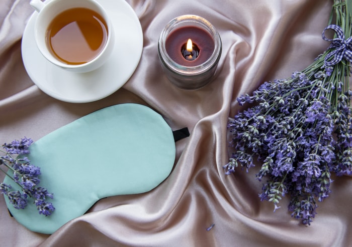 vue d'ensemble d'un plant de lavande étendu sur de la soie rose avec un masque de sommeil, un thé à la lavande et une bougie à la lavande allumée