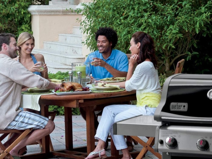 Quatre personnes mangeant à l'extérieur autour d'une table ronde à côté d'un barbecue Weber