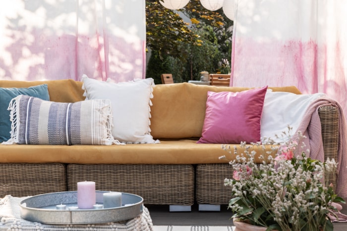 Coussins colorés sur un canapé en rotin sur la terrasse avec des fleurs, des rideaux peints et des bougies.