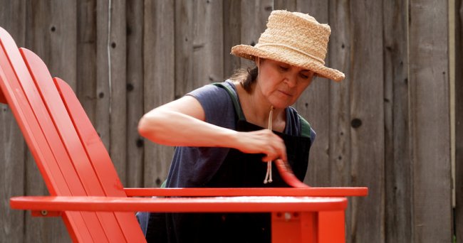 Une femme portant un chapeau à l'extérieur peint une chaise Adirondack en rouge.