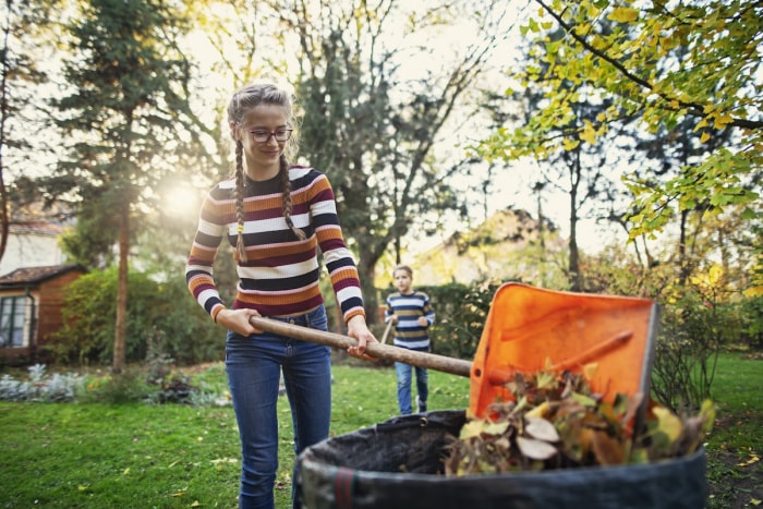 Une adolescente portant des lunettes et une chemise rayée jette des feuilles mortes dans un bac à compost