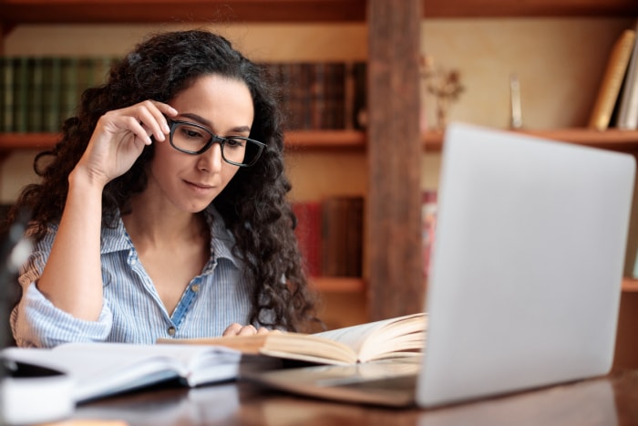 Jeune femme lisant un livre assise à un bureau touchant ses lunettes