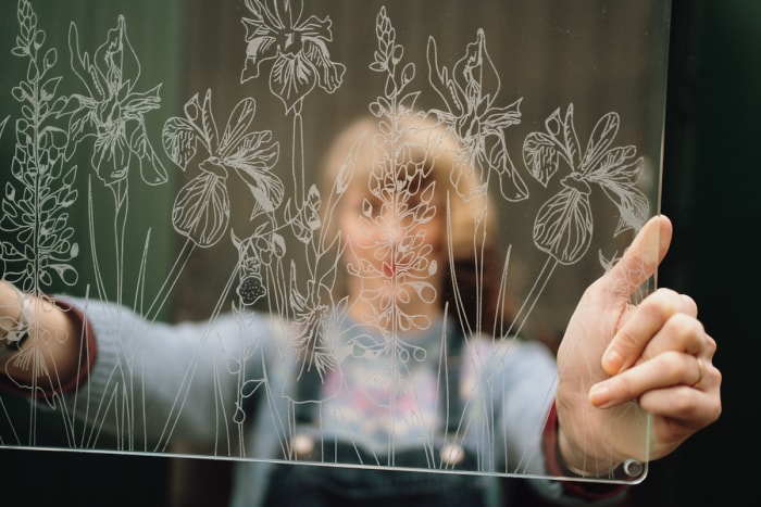 Prise de vue à travers un panneau de verre gravé avec un motif de prairie complexe. La femme qui le tient est floue de l'autre côté.