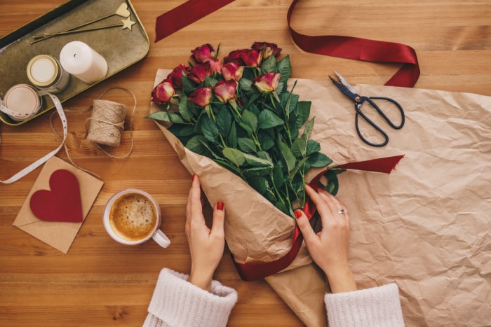 Vue de dessus des mains d'une femme créant un bouquet de fleurs de rose sur une table en bois avec des rubans et du papier