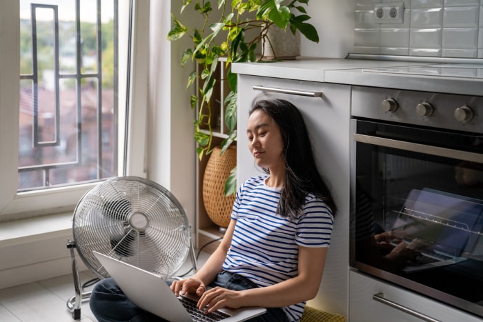 Jeune femme asiatique assise par terre dans la cuisine, tapant sur un ordinateur portable à côté d'un ventilateur électrique. 