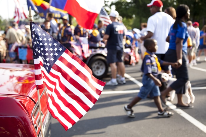 Drapeau américain flottant à l'arrière d'une voiture rouge vintage au premier plan d'une fête de quartier dans un parking