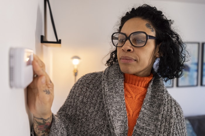 Une femme d'une quarantaine d'années portant des lunettes règle le thermostat du chauffage central de sa maison. Elle porte une couverture autour de ses épaules.