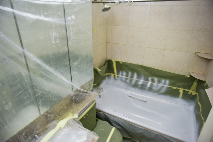 rénovation-de-salle-de-bain-avec-une-bâche-en-plastique-sur-le-lavabo-et-les-toilettes-tandis-que-la-baignoire-est-en-train-d'être-repeinte-en-blanc