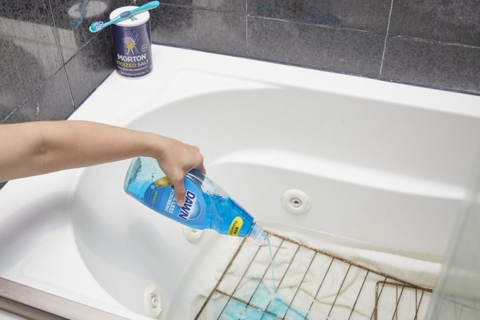 Une femme utilise du savon à vaisselle et du sel pour nettoyer les grilles du four dans une baignoire.