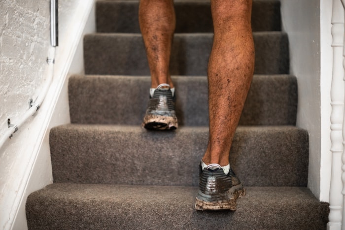 Une personne portant des chaussures de course boueuses monte un escalier recouvert de moquette grise.