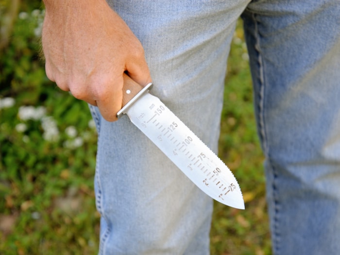 Une personne tenant le couteau hori hori Truly Garden à côté de sa jambe pour montrer sa jauge de profondeur.