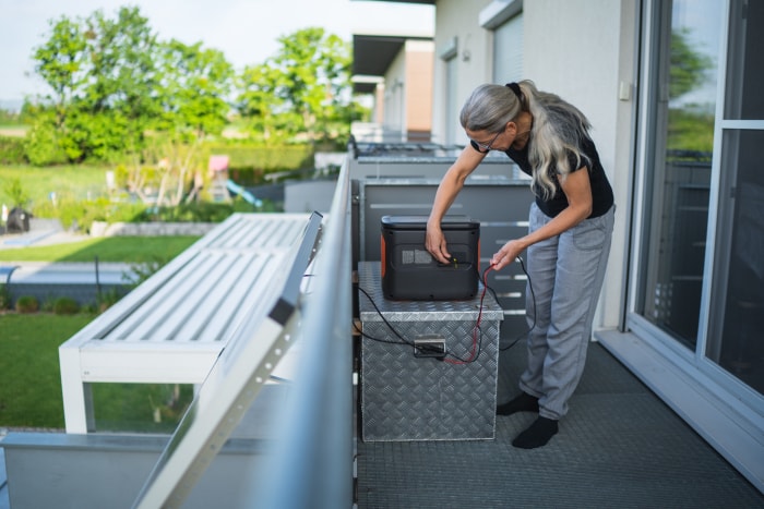 Une femme âgée branche le câble du panneau solaire sur un générateur portable sur le balcon de sa maison.