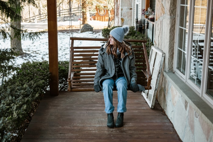 Une jeune femme en vêtements d'hiver est assise sur une balançoire de porche regardant la cour couverte de neige.