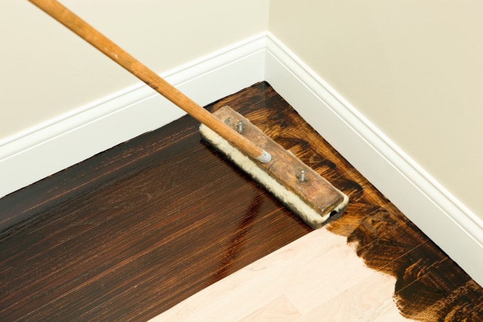 Un long pinceau applicateur est utilisé pour appliquer une teinture pour bois foncé sur un parquet poncé.